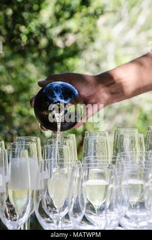Mans hand pours sparkling wine into glasses at outdoor event. Celebrating with champagne outside on sunny day. Stock Photo