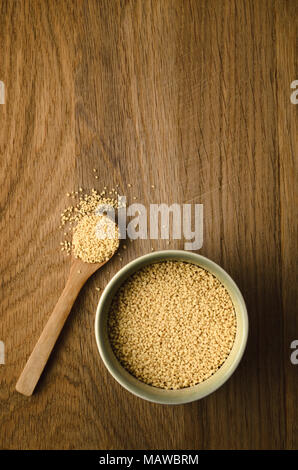 Overhead shot of couscous in a green ceramic bowl and upturned wooden spoon. Some grains spill on to oak wood surface background. Stock Photo