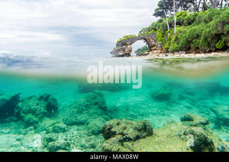 Split view over and under sea surface with lush tropical shore above waterline and corals with sand underwater. natural stone bridge on the shore. lif Stock Photo
