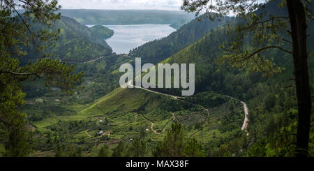 A thin road cut in to the volcanic hills around Lake Toba winds down to the lake viewed from west side through the surrounding trees. Sumatra, Indones Stock Photo