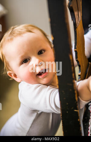 A 10 month old baby boy uses a chair to stand up, looking to camera. Stock Photo