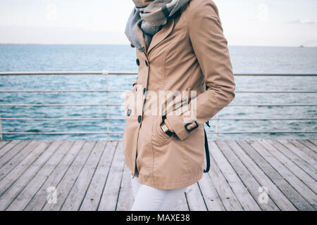 Fashionable girl standing on the waterfront. Cropped photo of slender young woman wearing beige trench coat, big scarf and gold wrist watches. Details Stock Photo