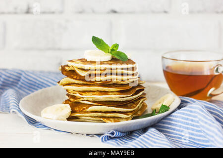 fresh baked pancakes with banana in white plate and cup of tea on blue linen on white background Stock Photo