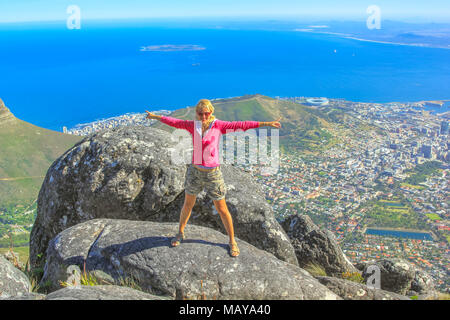 Young woman enjoying panoramic views of Cape Town from the top of Table Mountain National Park in Western Cape, South Africa. Happy female tourist with Port of Cape Town and Waterfront on background. Stock Photo