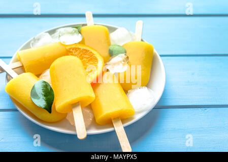 Orange popsicle on blue wooden background. Summer refreshing ices on stick. Stock Photo