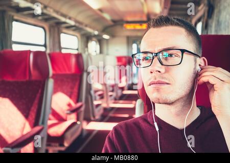 Rail transportation in sunny day. Thoughtful young man with eyeglasses listening to music while traveling by train. Stock Photo