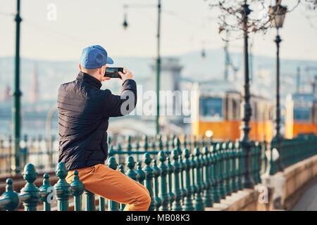 Young man (tourist) man sitting on the railing and taking picture with smart phone. Trams in Budapest, Hungary. Stock Photo