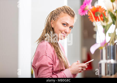 Smiling friendly young woman using a mobile phone indoors at home as she turns to smile at the camera viewed past colorful flowers Stock Photo