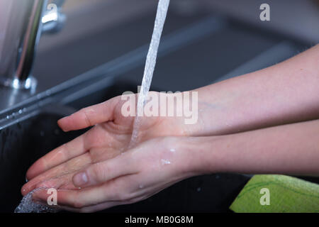 Young woman washing her hands in the kitchen under the running water from a tap on a stainless steel sink in a close up cropped view Stock Photo