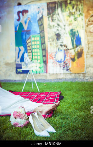 Woman resting her feet on a picnic blanket with colourful paintings in the background. Her shoes and peony bouquet are lying next to her feet Stock Photo