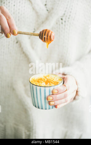 Healthy vegan turmeric latte or golden milk with honey in hands of woman wearing white sweater Stock Photo