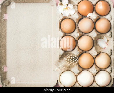 Easter holiday background. Flat-lay of gradient natural colored eggs in box with tender Spring almond blossom flower and feather over light pink backg Stock Photo