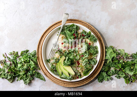 Vegan quinoa salad with kale, young beetroot leaves, garnet seeds, sliced avocado in white plate with ingredients above over grey texture background. Stock Photo