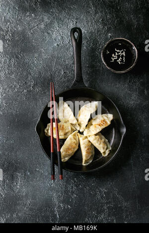 Asian dumplings Gyozas potstickers fried on cast-iron pan, served with chopsticks and bowl of soy sesame sauce over black texture background. Top view