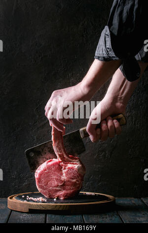 Man's hands cutting raw uncooked black angus beef tomahawk steak on bone by vintage butcher cleaver on round wooden slate cutting board over dark wood Stock Photo