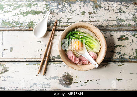 Asian dish udon noodles with egg yolk, sesame, mushrooms, boc choy, sliced sous vide cooked meat served in ceramic bowl with spoon and chopsticks over Stock Photo