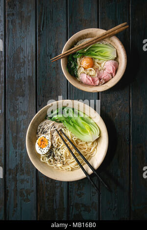 Set of Asian dish udon noodles with boiled egg, sesame, mushrooms, boc choy, sous vide pork meat served in ceramic bowls with chopsticks over dark woo Stock Photo