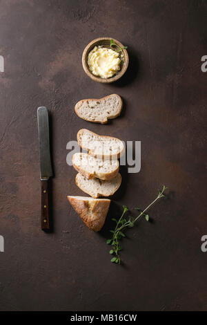 Loaf of fresh baked sliced artisan baguette bread with butter, herbs and vintage knife on wooden slate serving board over dark brown texture backgroun Stock Photo