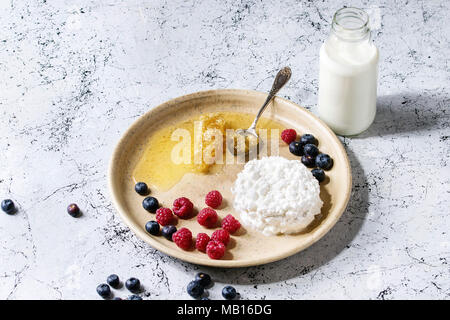 Ceramic plate of homemade cottage cheese served with blueberries, raspberries, bottle of milk and honeycombs over white marble texture table as backgr Stock Photo
