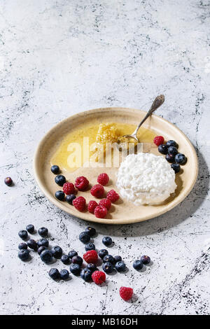 Ceramic plate of homemade cottage cheese served with blueberries, raspberries and honeycombs over white marble texture table as background. Stock Photo