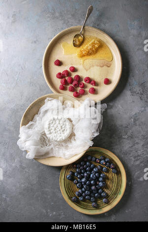 Fresh homemade cottage cheese in cheesecloth served in ceramic bowl with blueberries, raspberries and honeycombs over grey texture background. Top vie Stock Photo