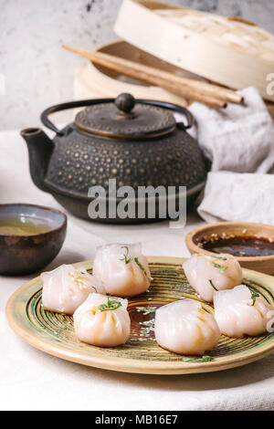 Asian steam potstickers dumplings stuffed by shrimps, served on ceramic plate with soy sesame sauce, chopsticks, teapot, tea, bamboo steamer over kitc Stock Photo