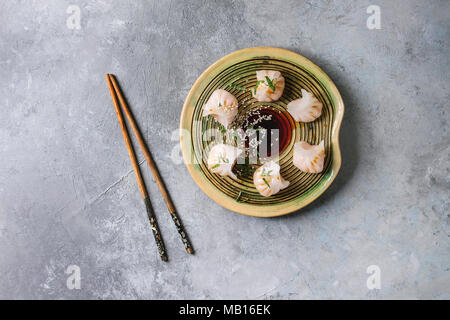 Asian steam potstickers dumplings stuffed by shrimps, served on ceramic plate with soy sesame sauce and chopsticks over grey texture background. Top v Stock Photo