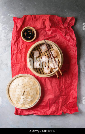 Asian steam potstickers dumplings stuffed by shrimps, served in opened bamboo steamer with soy sesame sauce and chopsticks on crumpled red paper over Stock Photo