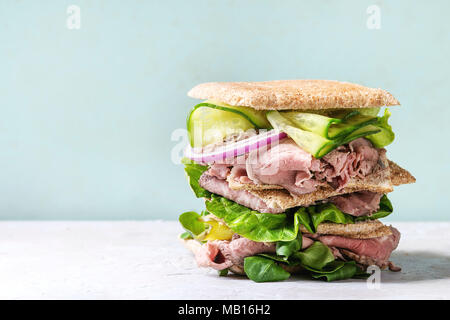 Beef and vegetables sandwiches with sliced meat, cucumber, green salad, rye whole grain bread in stack over grey green pin-up style background. Stock Photo