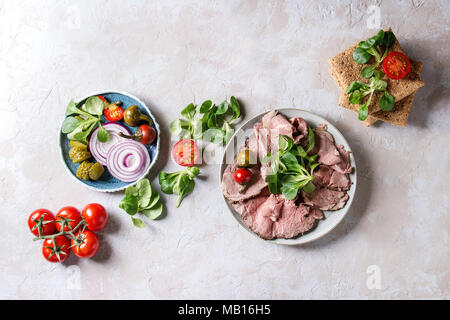 Ingredients for making sandwiches. Sliced beef meat, rye whole graine bread, green salad, tomatoes, pickled cucumber, onion on blue plates over grey t Stock Photo