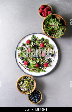 Green vegan salad with berries strawberry, blueberry, sprouts, young beetroot leaves on ceramic plate. Ingredients in wooden bowls above over grey con Stock Photo