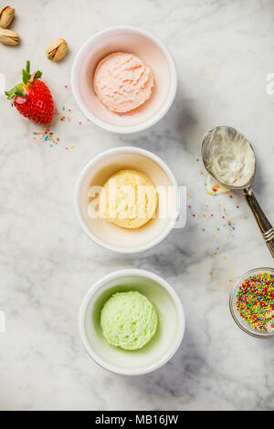 Top view of ice cream in white bowls and fresh ingredients on white marble background. Pink (strawberry), yellow (mango or banana) and green (lime, gr Stock Photo