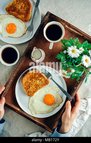 A woman holding a wooden tray with an easy French toast served with a fried egg on the side on a white plate, a cup of coffee, milk and spring flowers Stock Photo