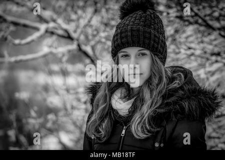 Closeup portrait of pretty teen girl at winter, black and white. Beautiful Caucasian teenage girl in black coat, blurred snowy forest in background. Stock Photo