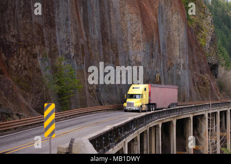 Yellow bonnet American big rig semi truck carry cargo in long roomy bulk semi trailer on winding dangerous road with rock wall and bridge on the slope Stock Photo