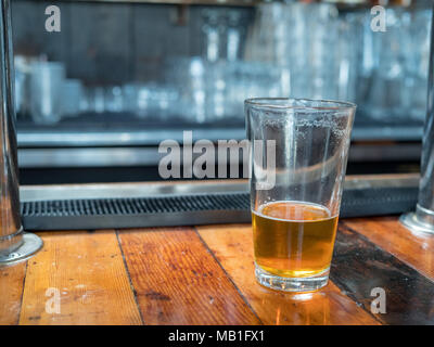 Almost empty pint glass of beer sitting on woodencountertop for last call. Bar in background Stock Photo