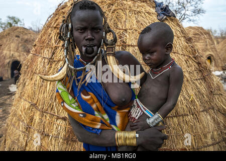 Omorate Ethiopia - January 24, 2018: Woman of the Mursi tribe with traditional jewelry and her son with the typical houses of the Mursi in Ethiopia. Stock Photo