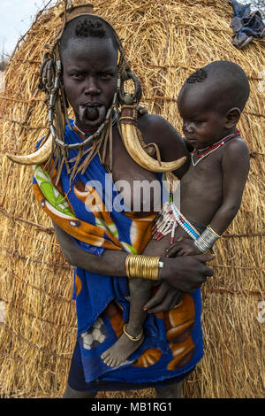 Omorate Ethiopia - January 24, 2018: Woman of the Mursi tribe with traditional jewelry and her son with the typical houses of the Mursi in Ethiopia. Stock Photo