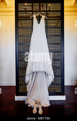 The front view of wedding dress hanging on the wall with shoes on the floor during getting ready, tungsten light in background. Stock Photo