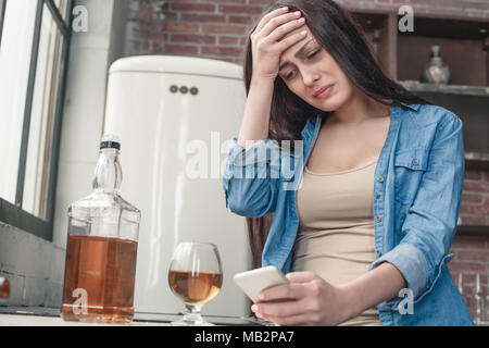 Young female alcoholic social problems standing holding phone looking at pictures memories crying desperate touching head bottom view Stock Photo