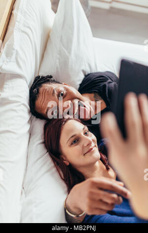 Top view of multiracial couple lying together on bed and taking selfie. Man sticking out tongue with woman taking selfie using mobile phone. Stock Photo