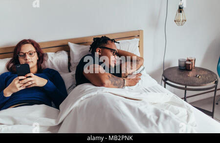 Young multiracial couple using their mobile phones in bed ignoring each other. Man and woman busy with their cell phones in bed. Stock Photo