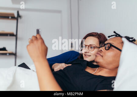 Interracial couple lying together on a bed and using a mobile phone. Young man and woman in bed looking at smart phone. Stock Photo