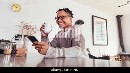 Happy young african man sitting at table using mobile phone and drinking coffee. Man relaxing at home reading text message on his cell phone in mornin Stock Photo