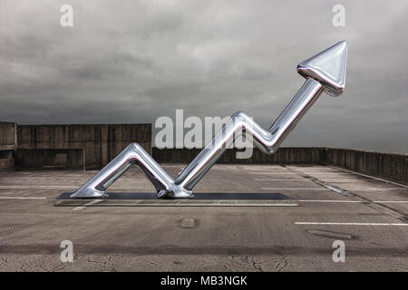 A shiny arrow made from chrome or steel sits on a car park pointing upwards Stock Photo