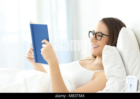 Happy woman wearing eyeglasses reading a book lying on the bed at home or hotel room Stock Photo