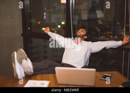 relaxed confident young businessman sitting with legs on desk at office work late overtime at night Table footrest Stock Photo