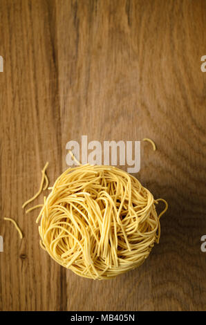 Overhead shot of egg noodles nest stack on wooden surface with broken noodle fragments scattered below. Copy space above. Stock Photo