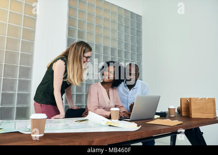 Group of diverse office colleagues discussing blueprints and working on a laptop at a desk in a modern office Stock Photo