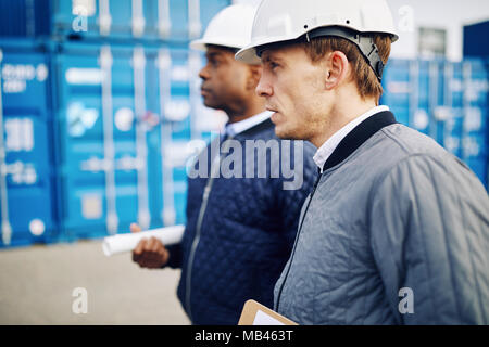 Two engineers wearing hardhats standing together by freight containers on a large commercial shipping dock Stock Photo